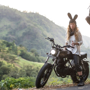 A person wearing a black bunny beanie on a motorcycle with a green natural background