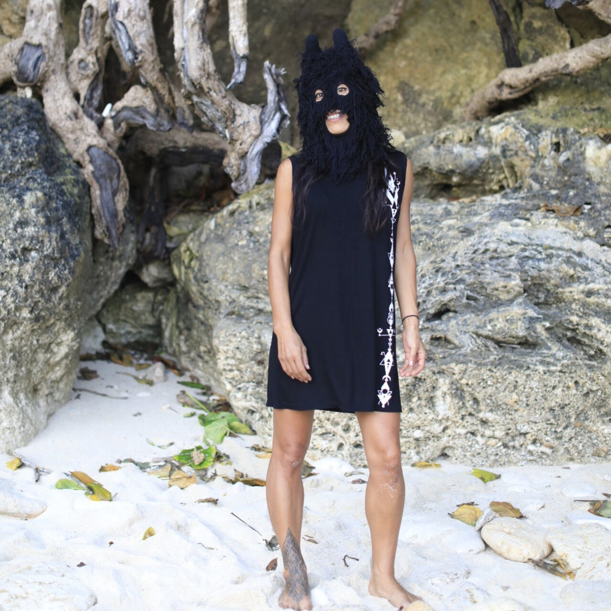 Person wearing a black dress with white patterns and a black mask on a rocky beach.