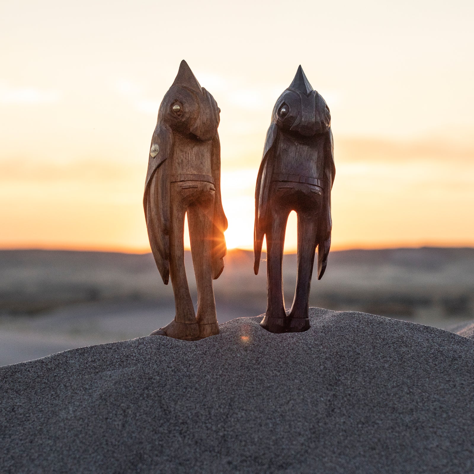 Two wooden bird figures standing on sand with a sunset in the background