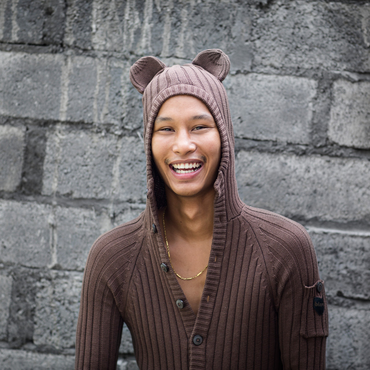 Person wearing a brown knit hoodie with bear ears against a brick wall.