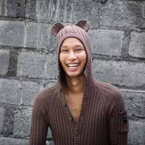 Person wearing a brown knit hoodie with bear ears against a brick wall.