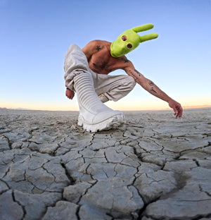 A man wearing a lime green balaclava with horns and white pants and futuristic shoes. 