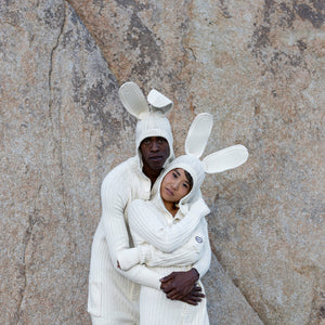 Two people in an embrace wearing white bunny onesies standing in front of a rocky background 