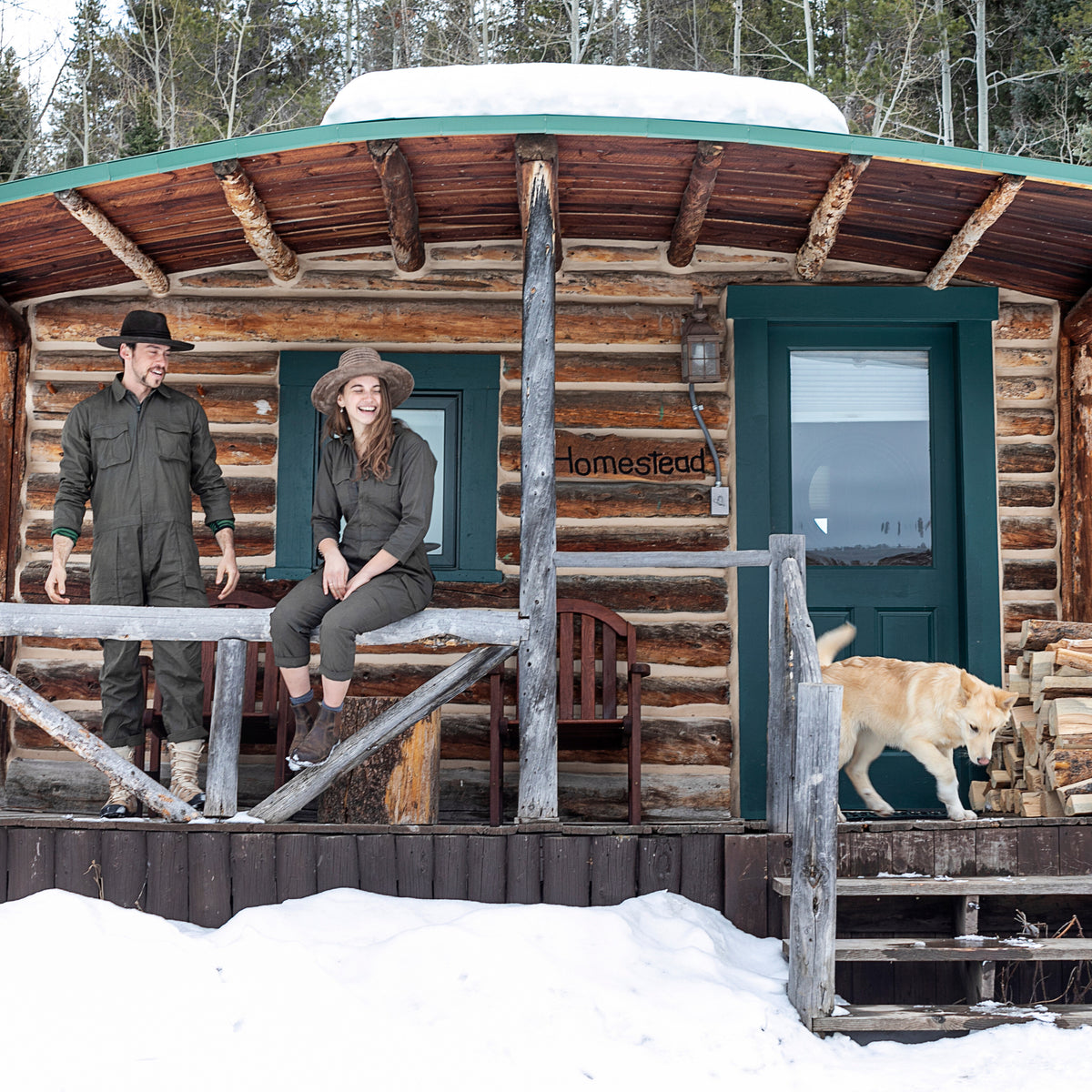 Two people wearing green jumpsuits and hats smiling with a dog on the porch of a wooden cabin in the snow