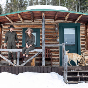 Two people wearing green jumpsuits and hats smiling with a dog on the porch of a wooden cabin in the snow