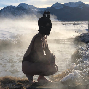 Person sitting by a hot spring wearing a balaclava mask with ears with snow-covered mountains in the background