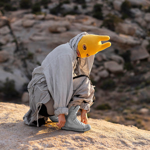 Person kneeling down with a yellow mask and gray outfit on a rocky landscape