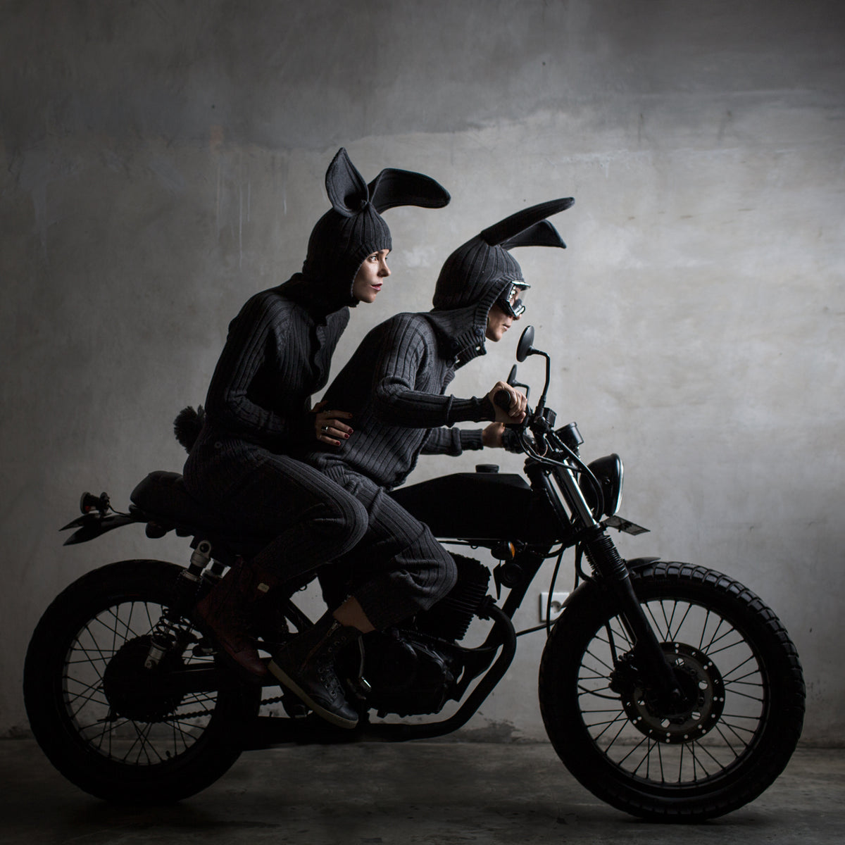 Two people in dark clothing and bunny ears riding a motorcycle against a gray background