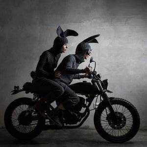 Two people in dark clothing and bunny ears riding a motorcycle against a gray background