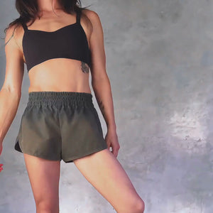 Person wearing a black cropped tank and olive green athletic shorts spinning against a gray background 