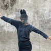 Person in an indigo outfit and indigo balaclava mask with pointed ears dancing against a gray textured background 