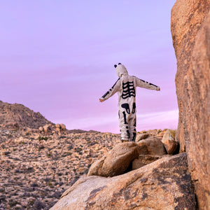 Person wearing a skeleton onesie with their arms outstretched standing on a rock with a rocky terrain and clear skies behind them