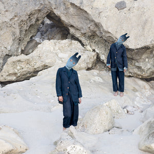 Two people wearing blue masks and outfits with their heads tilted sideways against a rocky background