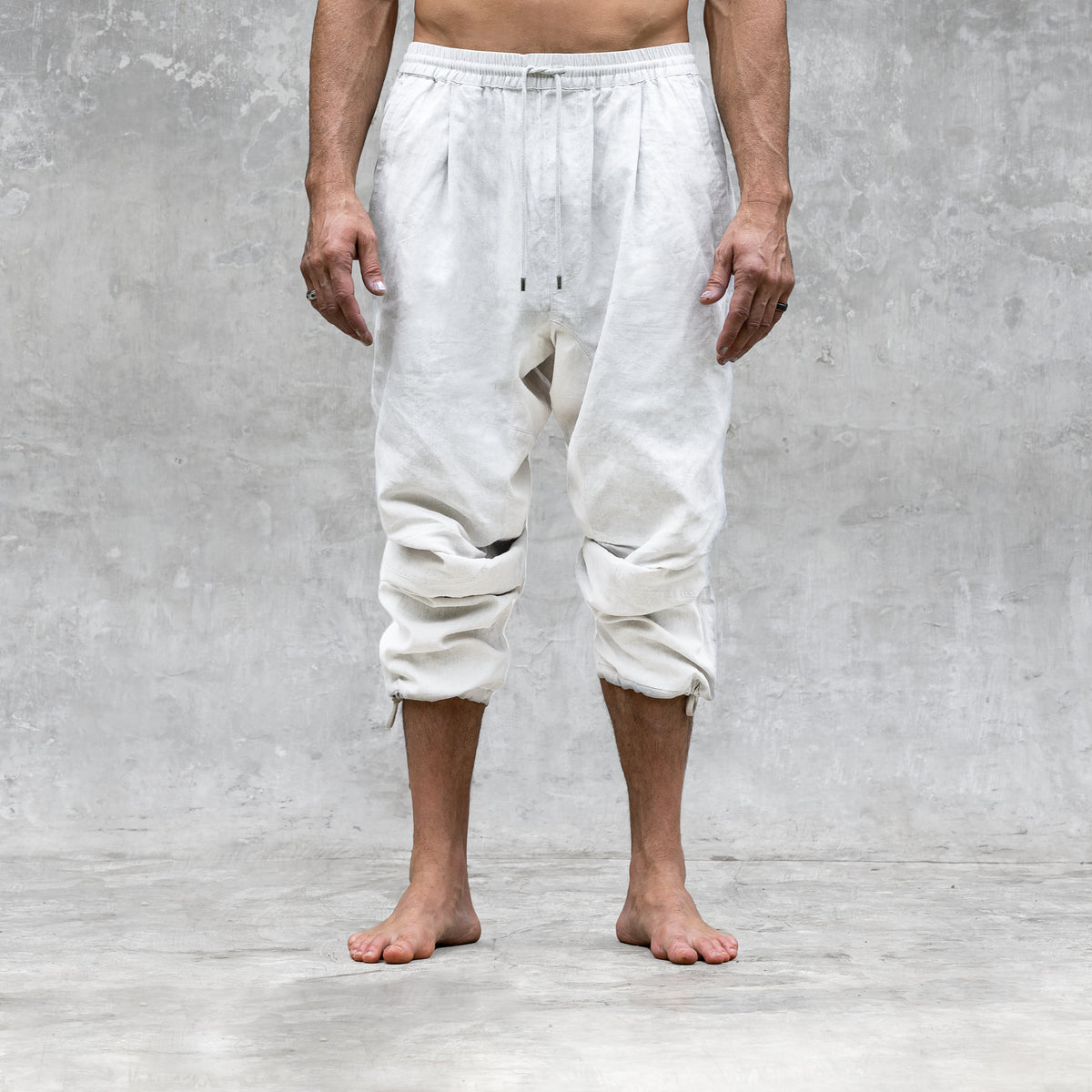 A man standing barefoot and shirtless from the feet to the waist facing front wearing a pair of white parachute style linen pants. 