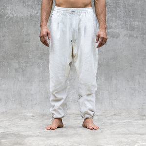 A man standing barefoot and shirtless from the feet to the waist facing front wearing a pair of white parachute style linen pants. 