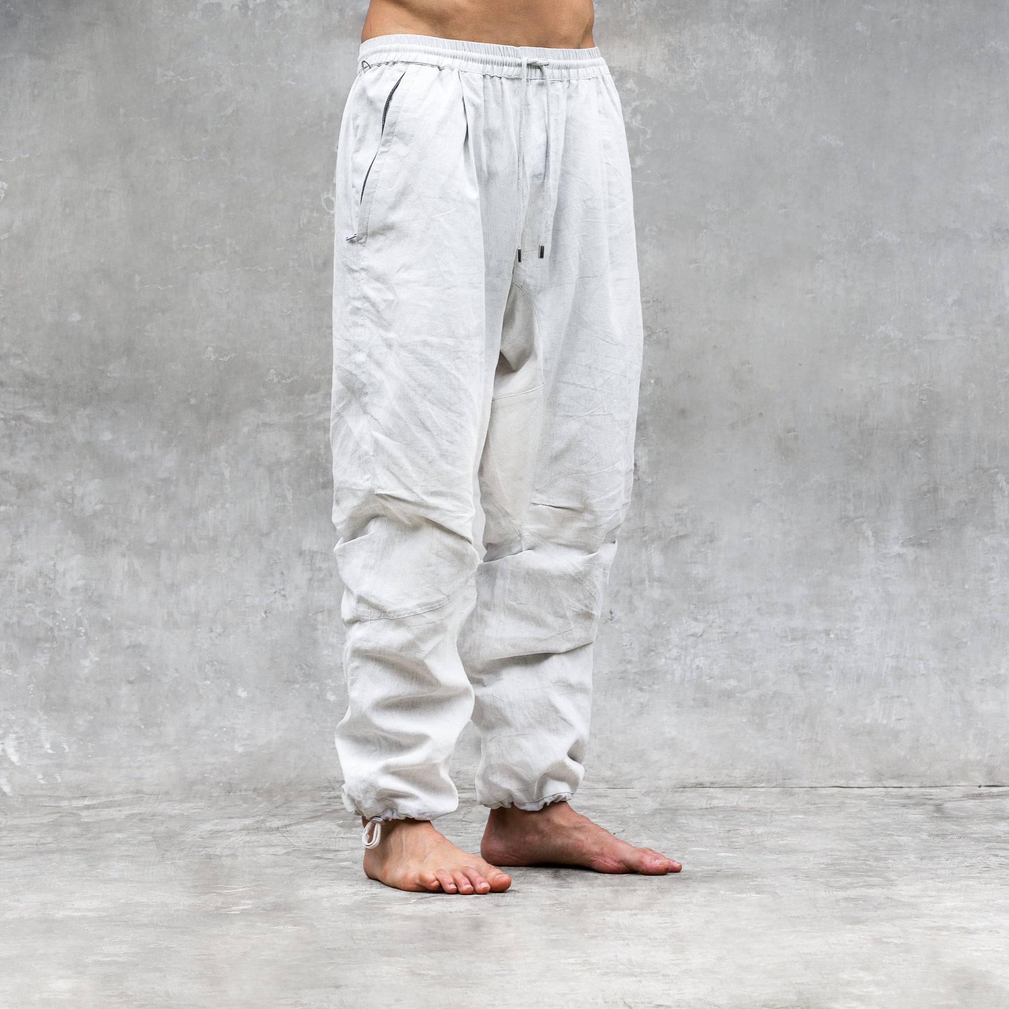 A man standing barefoot wearing a pair of white parachute style linen pants. 