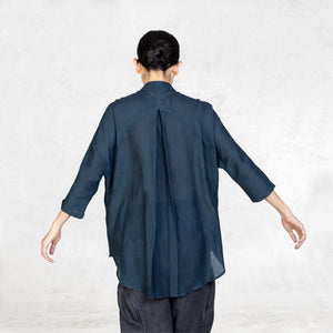 A woman standing from the waist up facing back with her arms lifted to the side wearing dark indigo pants and an Indigo Blue 3/4 sleeve cotton silk button up with an asian style collar