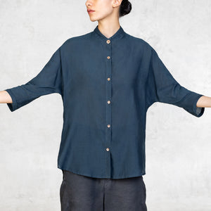 A woman standing from the waist up facing front with her arms lifted to the side wearing dark indigo pants and an Indigo Blue 3/4 sleeve cotton silk button up with an asian style collar