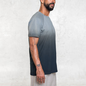 Man standing from the waist up facing sideways wearing a short sleeve cotton tee shirt that fades from indigo blue at the hem toward light blue at the shoulders