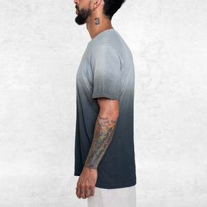 Man standing from the waist up facing sideways wearing a short sleeve cotton tee shirt that fades from indigo blue at the hem toward light blue at the shoulders