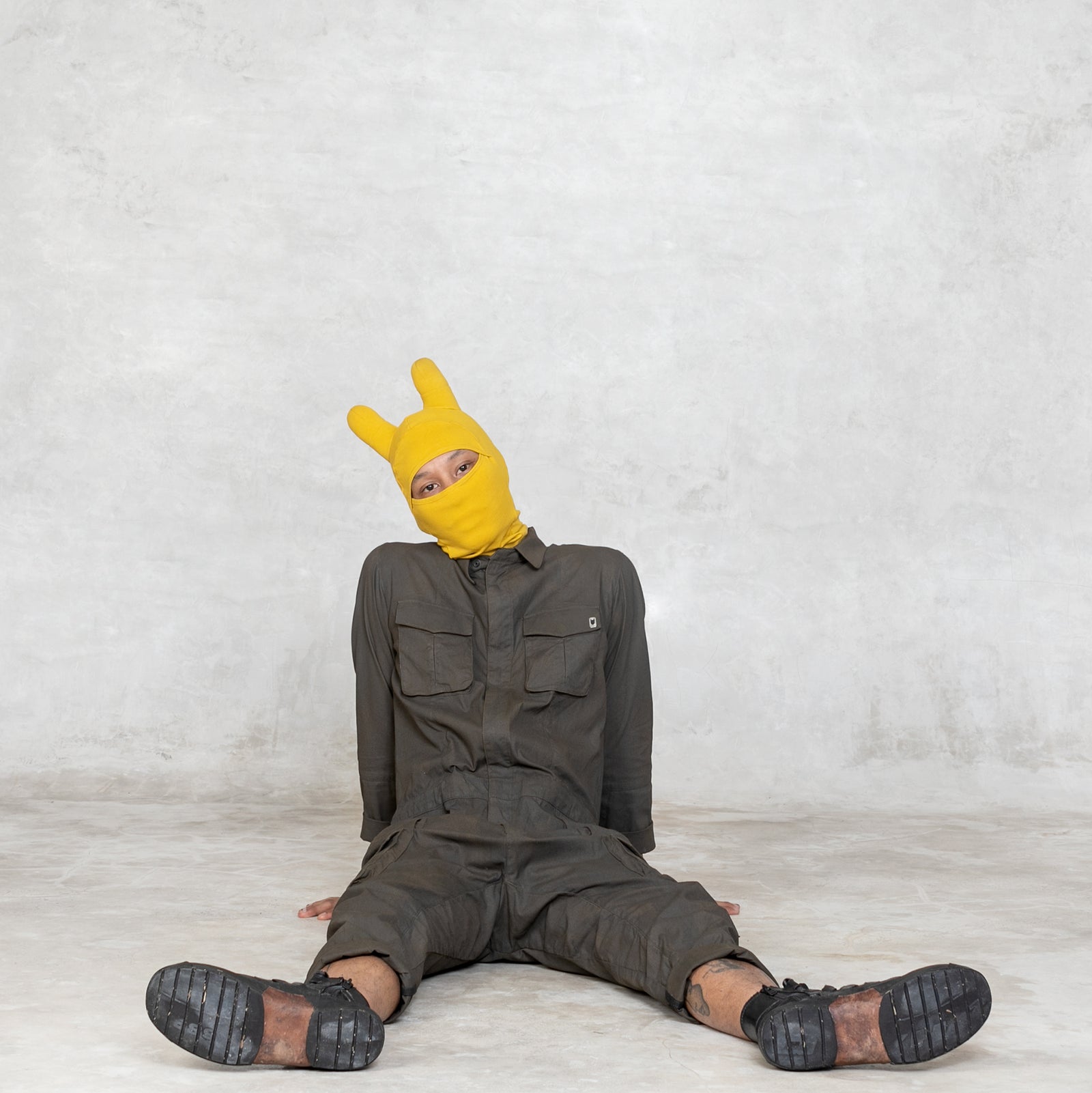 Person wearing a yellow balaclava mask with rounded ears, an olive green jumpsuit, and black and brown shoes sitting on a gray surface 