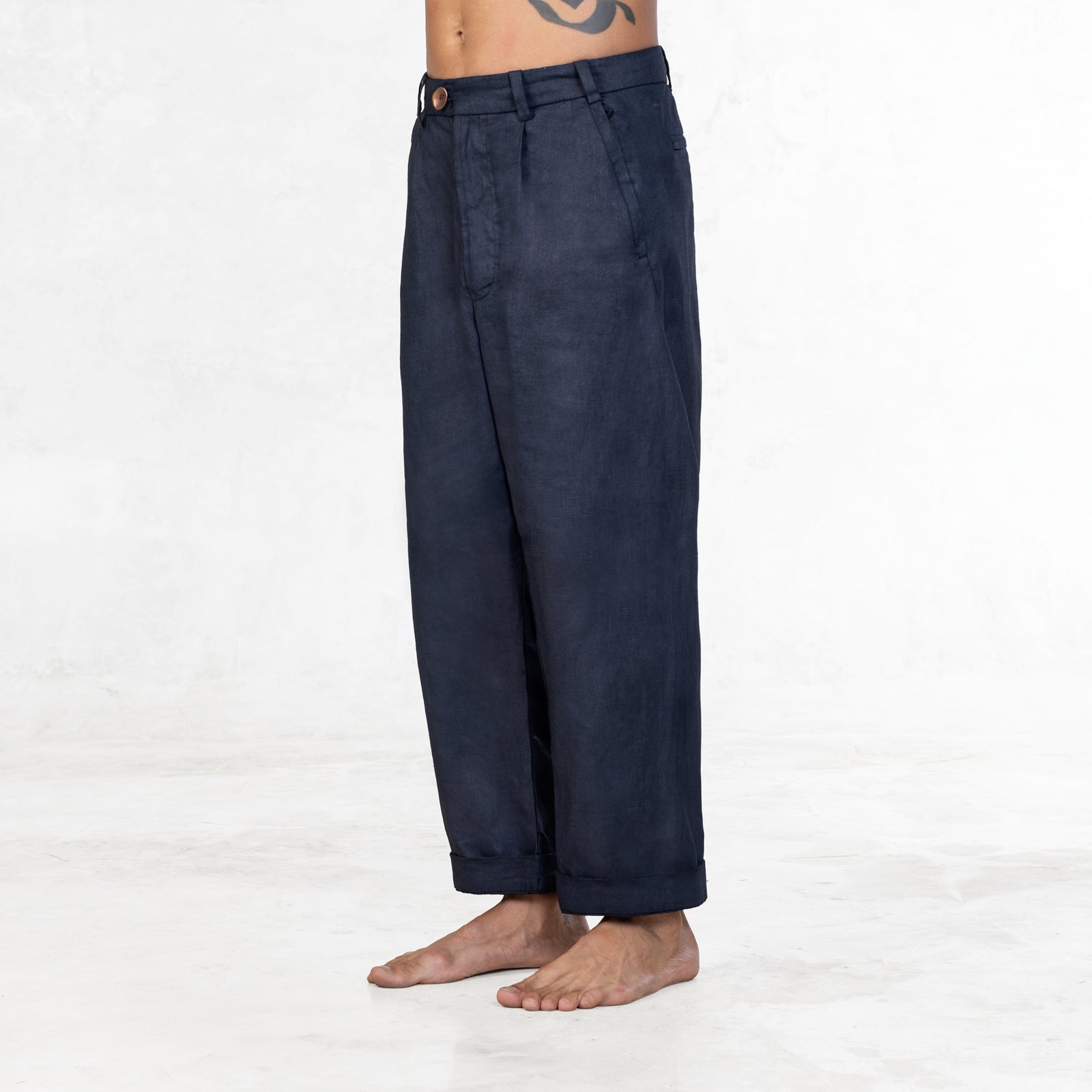 A shirtless man standing facing slightly side from the waist down wearing Indigo blue linen pants and blue suede military style boots