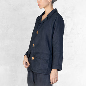 A woman standing facing side from the thighs up wearing an Indigo Blue Linen suit. A 3 button button-up jacket with 3/4 sleeves.
