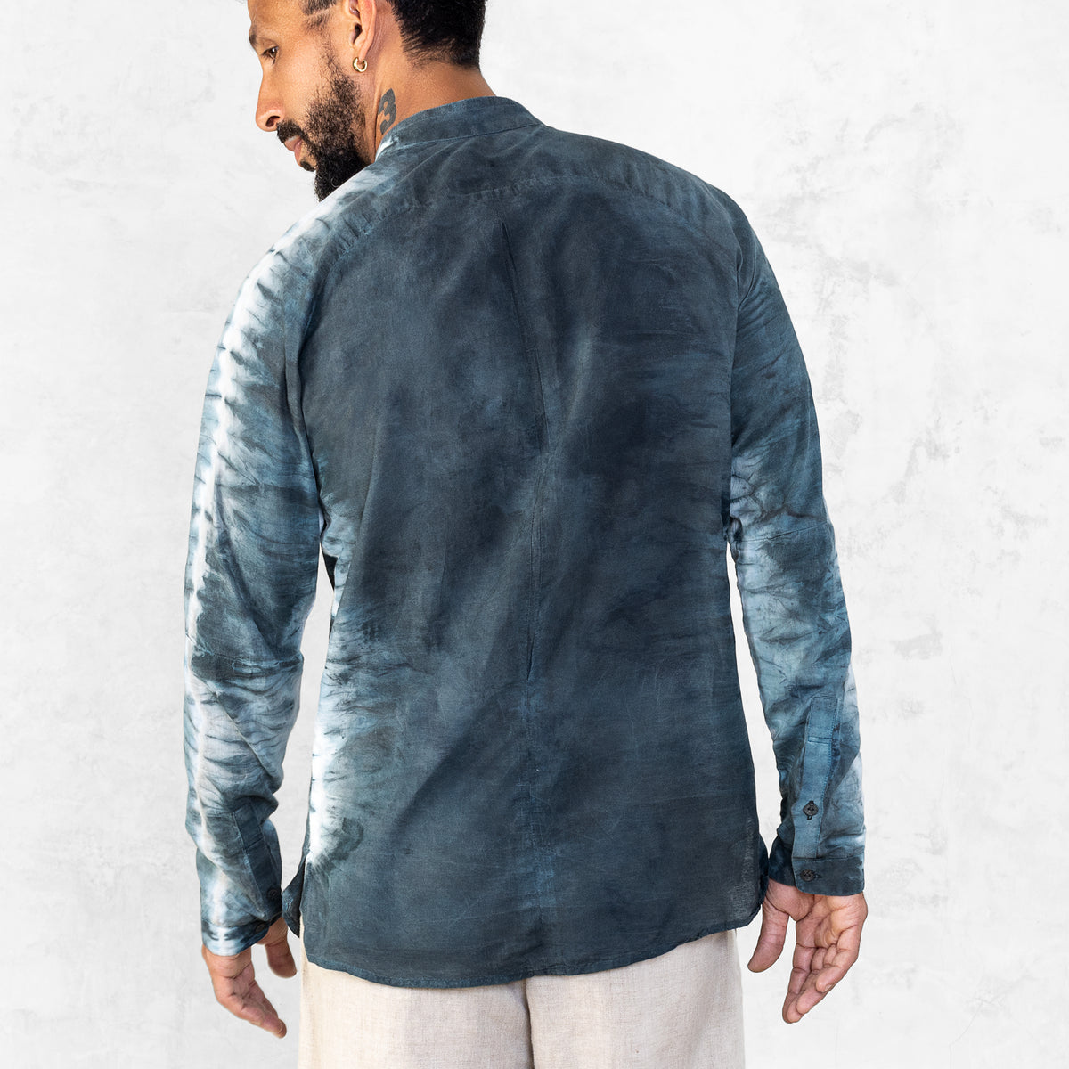A man standing from the waist up facing back wearing white pants and an Indigo Blue long sleeve cotton silk button up with tie dyed arms