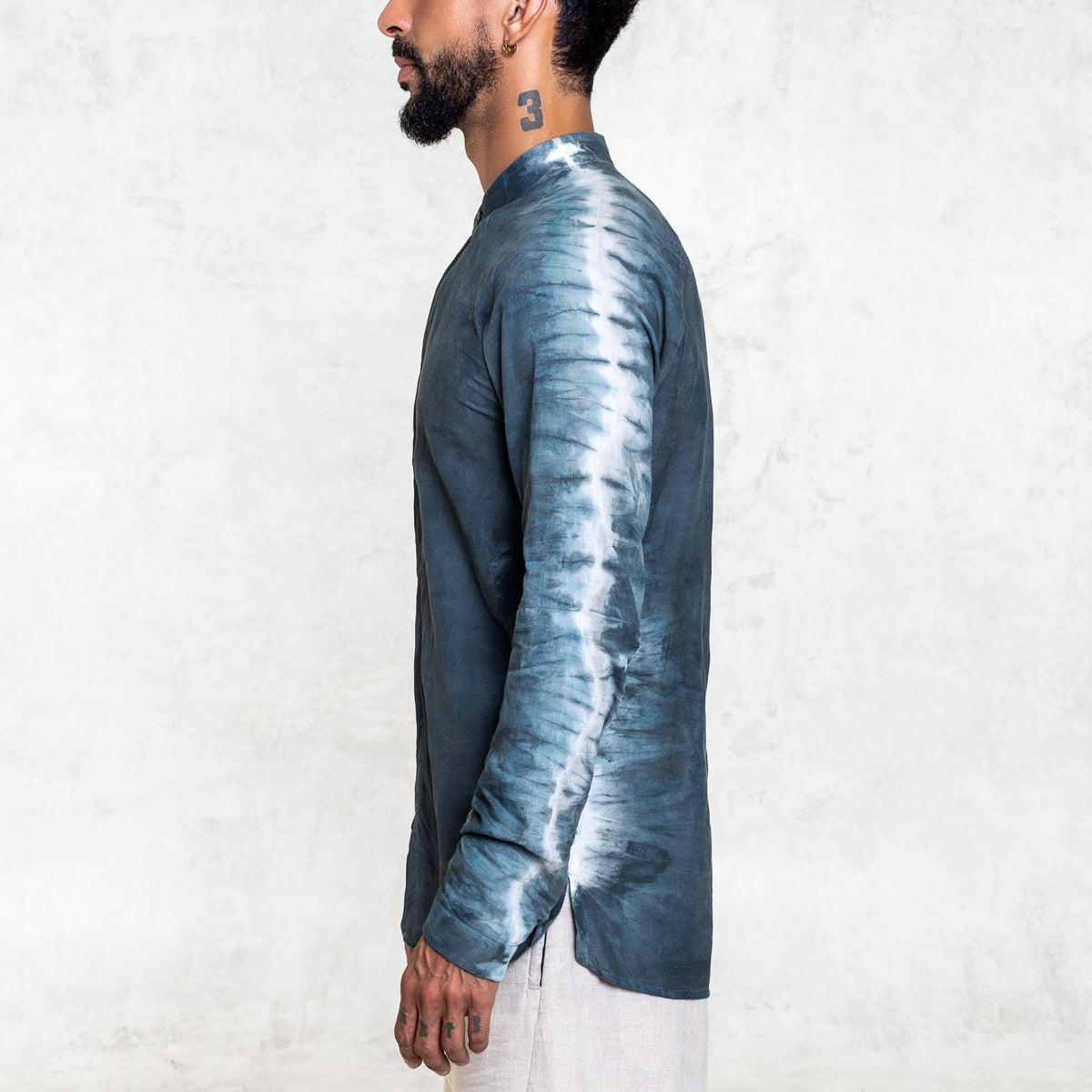 A man standing from the waist up facing to the side wearing white pants and an Indigo Blue long sleeve cotton silk button up with tie dyed arms