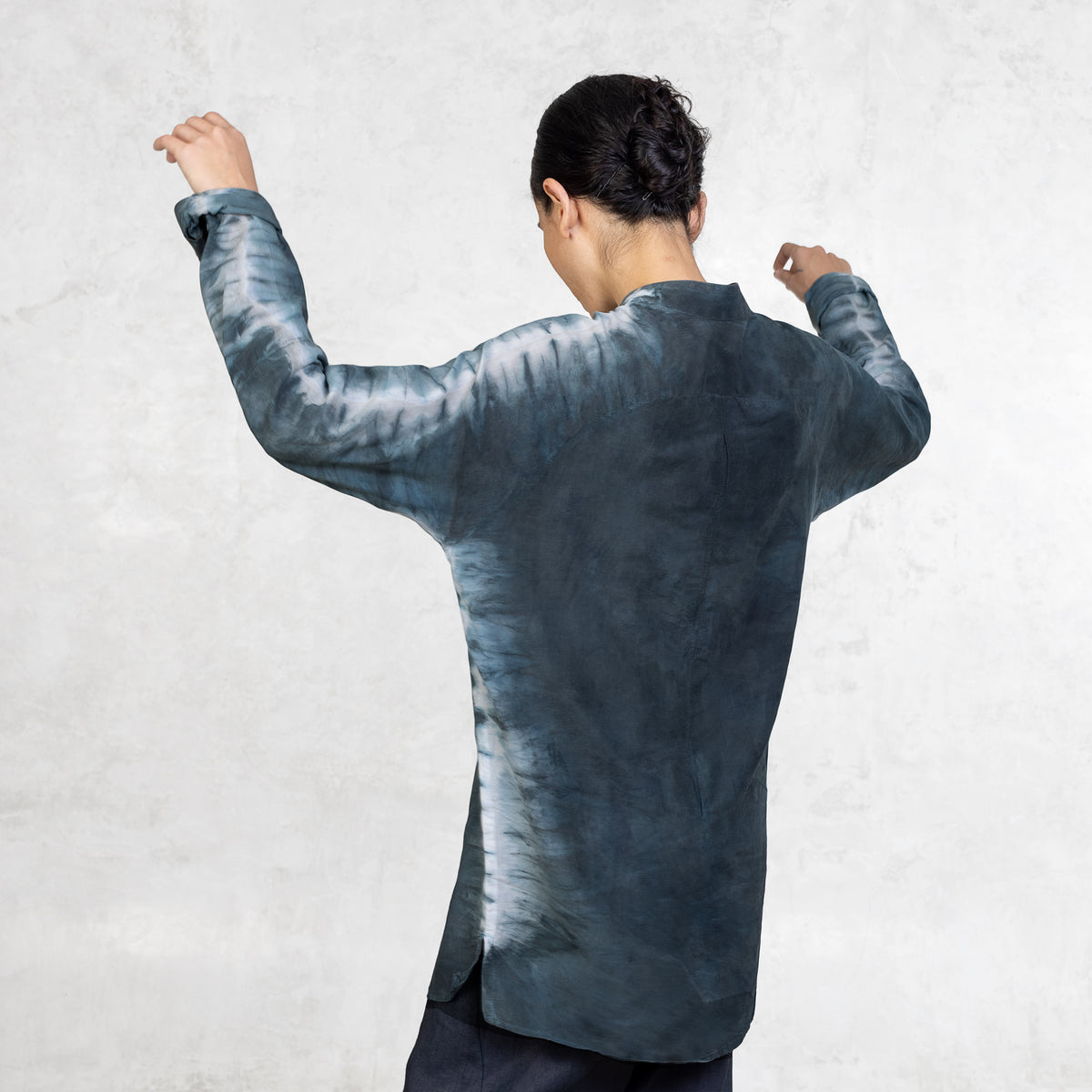 A woman standing from the waist up facing back with her arms lifted wearing dark indigo pants and a light Indigo Blue long sleeve cotton silk button up with tie dyed arms