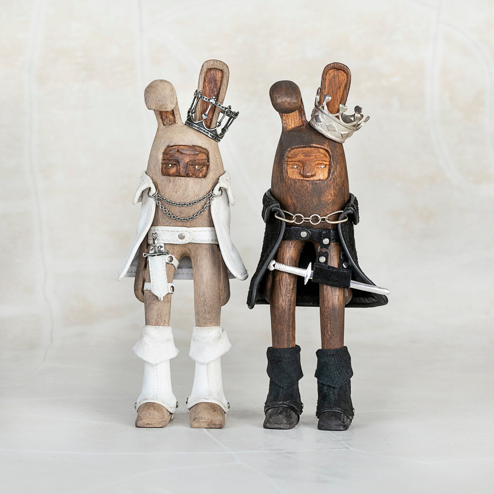 Two wooden bunny rabbit figurines wearing silver crowns, leather capes, and leather boots with silver daggers and other silver accents, standing side by side against a white background