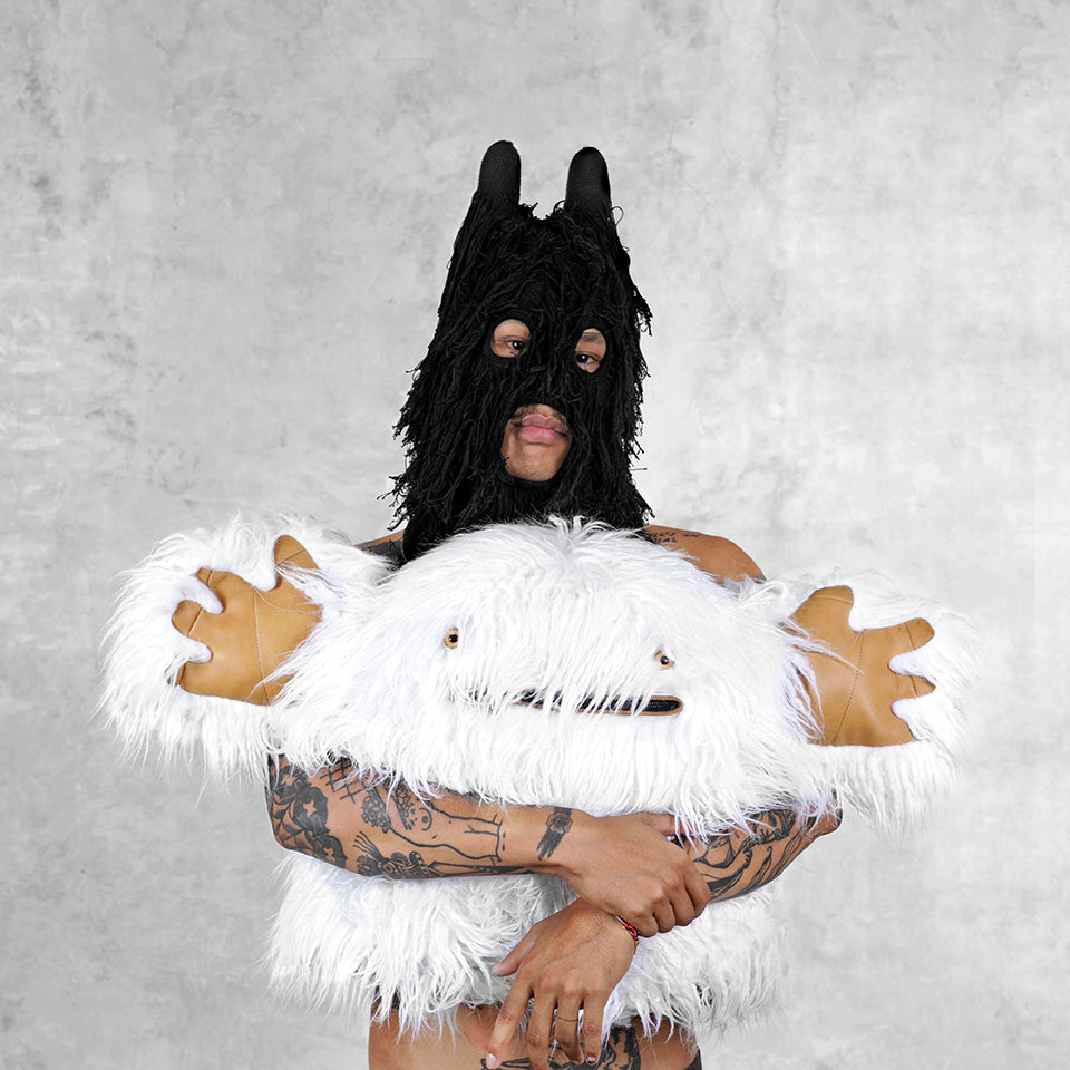 Tattooed person wearing a black stringy mask with rounded ears holding a white hairy creature plush against a light gray textured background 