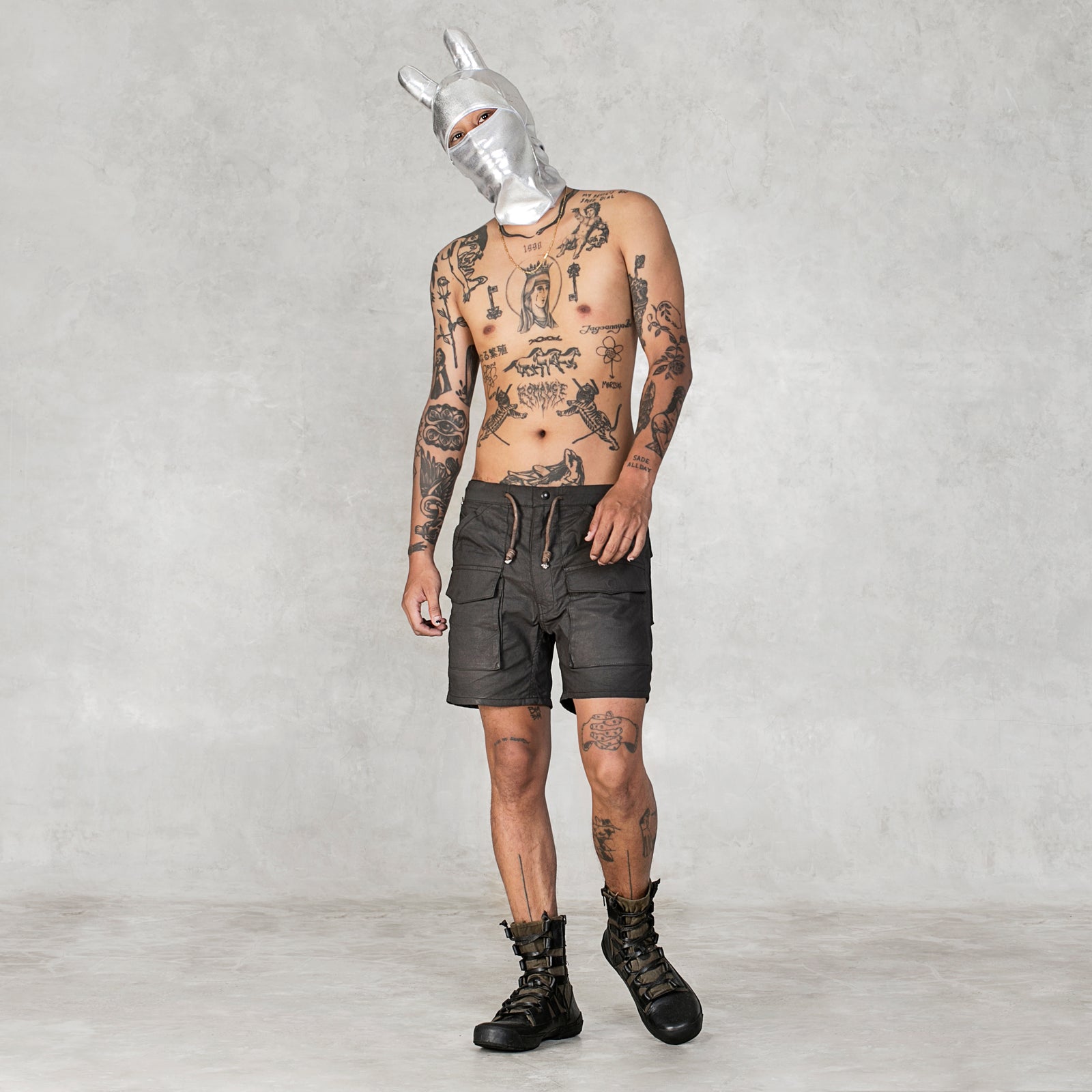 Shirtless tattooed person wearing a silver balaclava mask with rounded ears, black cargo style shirts, and black boots standing in front of a light gray background 