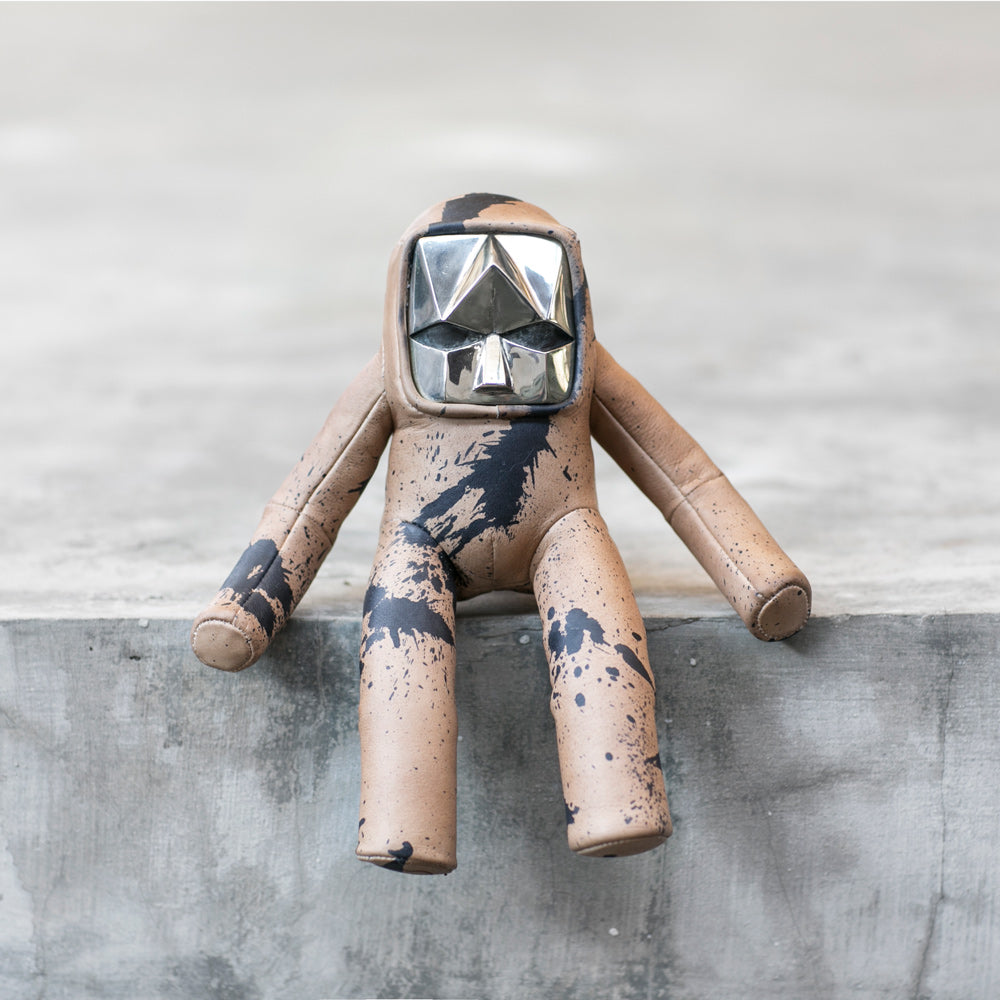 A toy figure with a beige and black splattered design and a silver geometric headpiece sitting on the edge of a gray textured ledge