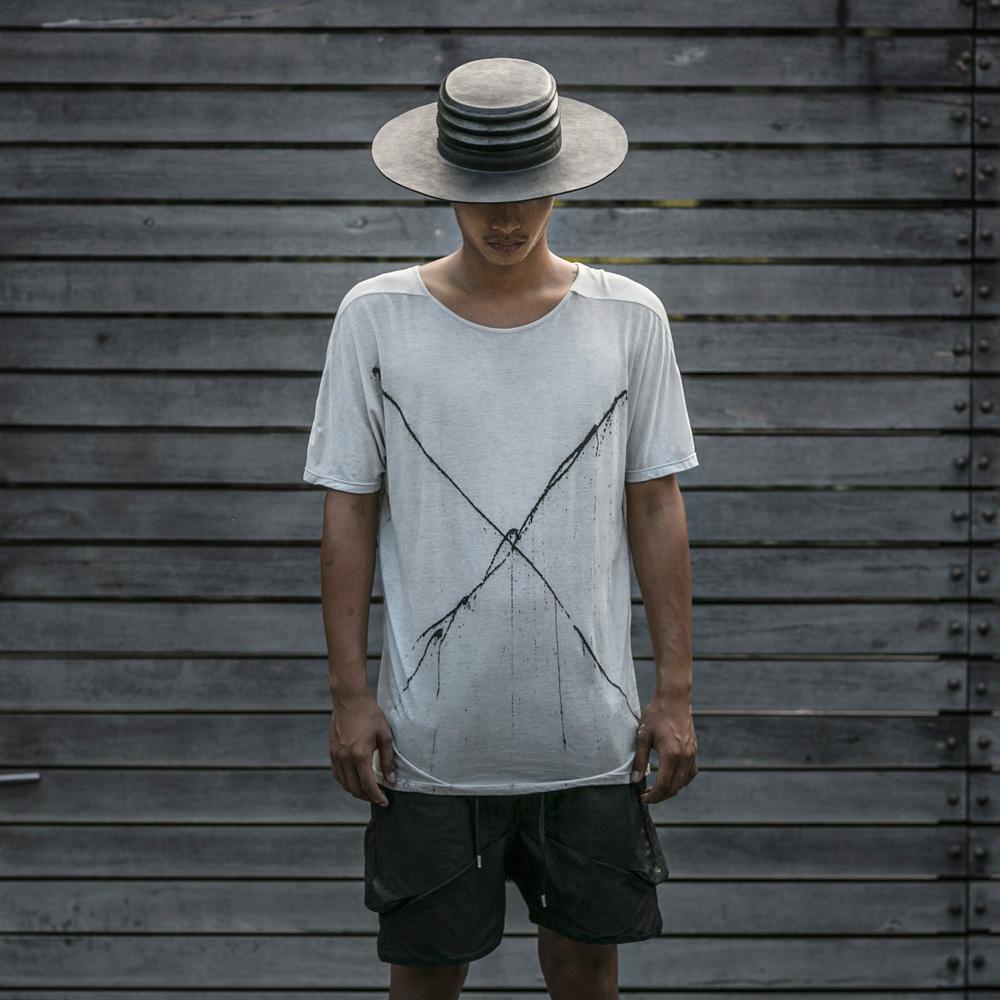 A person wearing a Blamo Verge Hat Black with a white graphic tee and black drawstring shorts standing in front of a wooden plank background 