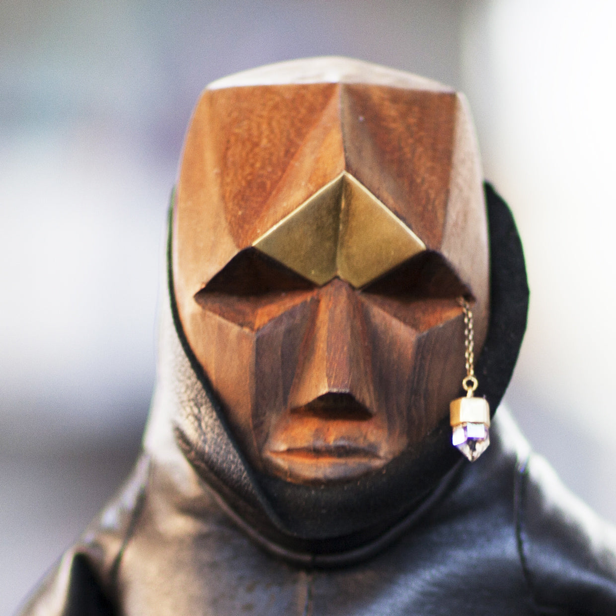 Closeup of a wooden, leather, and brass figurine with a geometric head and a quartz hanging from a brass chain near its eye against a blurred background